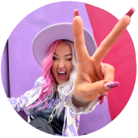 Person with pink hair smiling and making a peace sign with their hand in front of colorful background.
