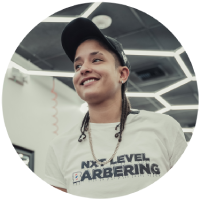 Smiling barber wearing black cap and white shirt with 'Nxt Level Barbering' logo indoors.