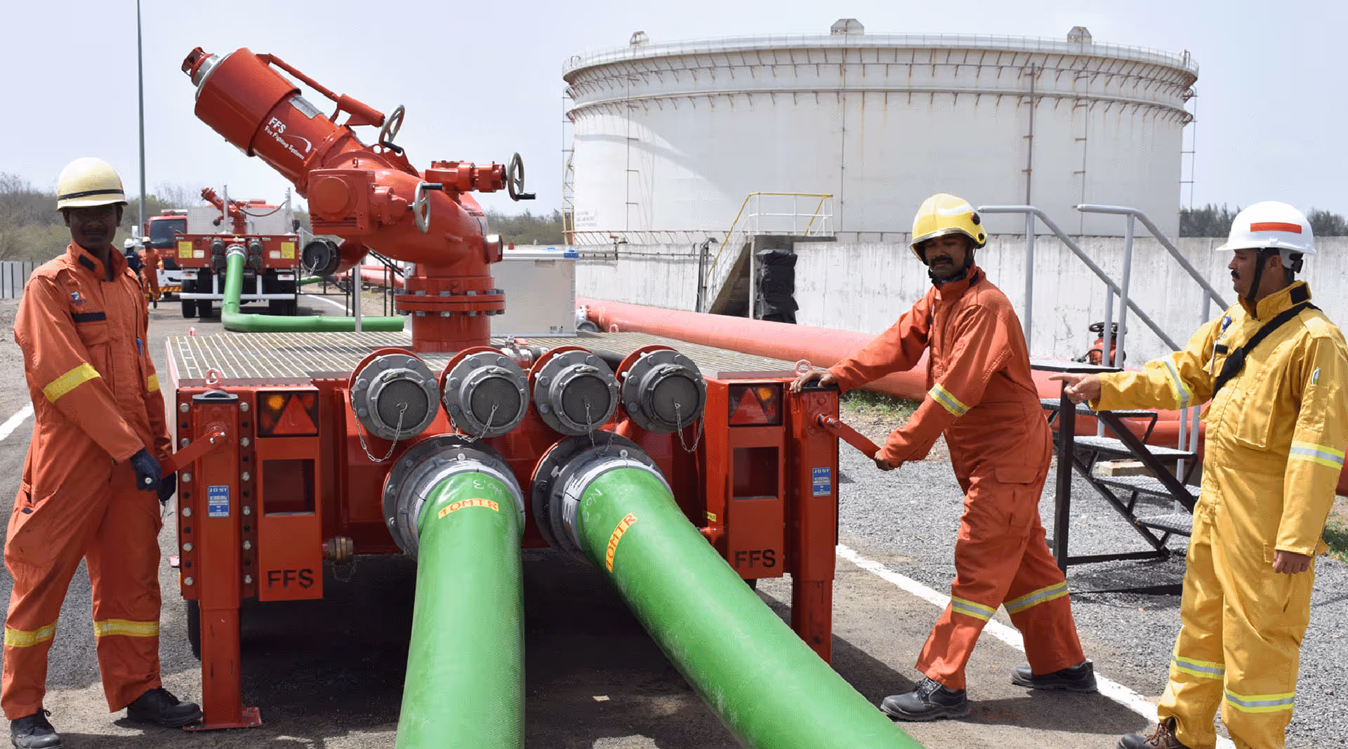 Three workers in safety helmets and protective suits operate large red firefighting equipment attached to green hoses outside an industrial facility.