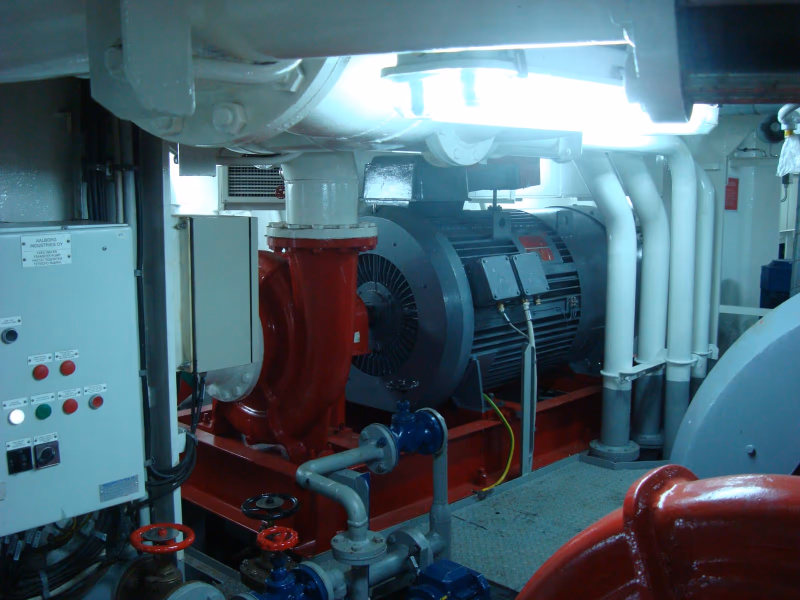 Industrial electric motor installed with red and white pipes and a control panel nearby in a machinery room.