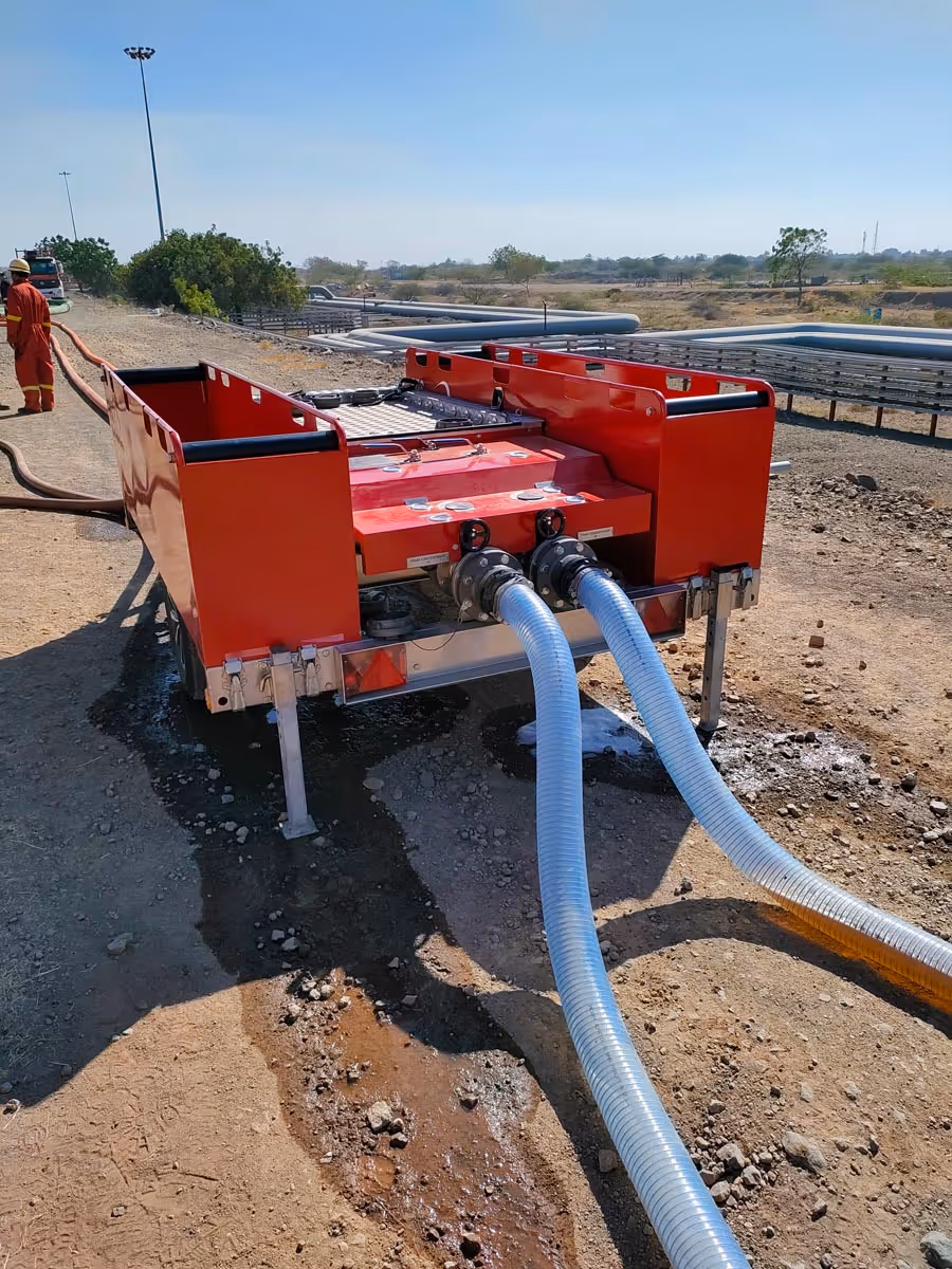 Red industrial water pump connected to two large transparent hoses on a dirt road with a worker wearing orange safety gear standing nearby.