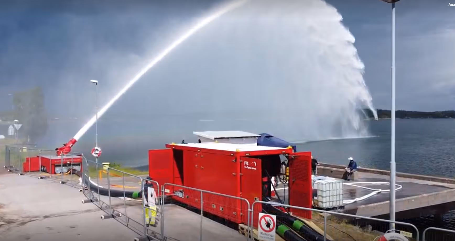 Red fire pump machine spraying a large arc of water into a lake from a dockside.