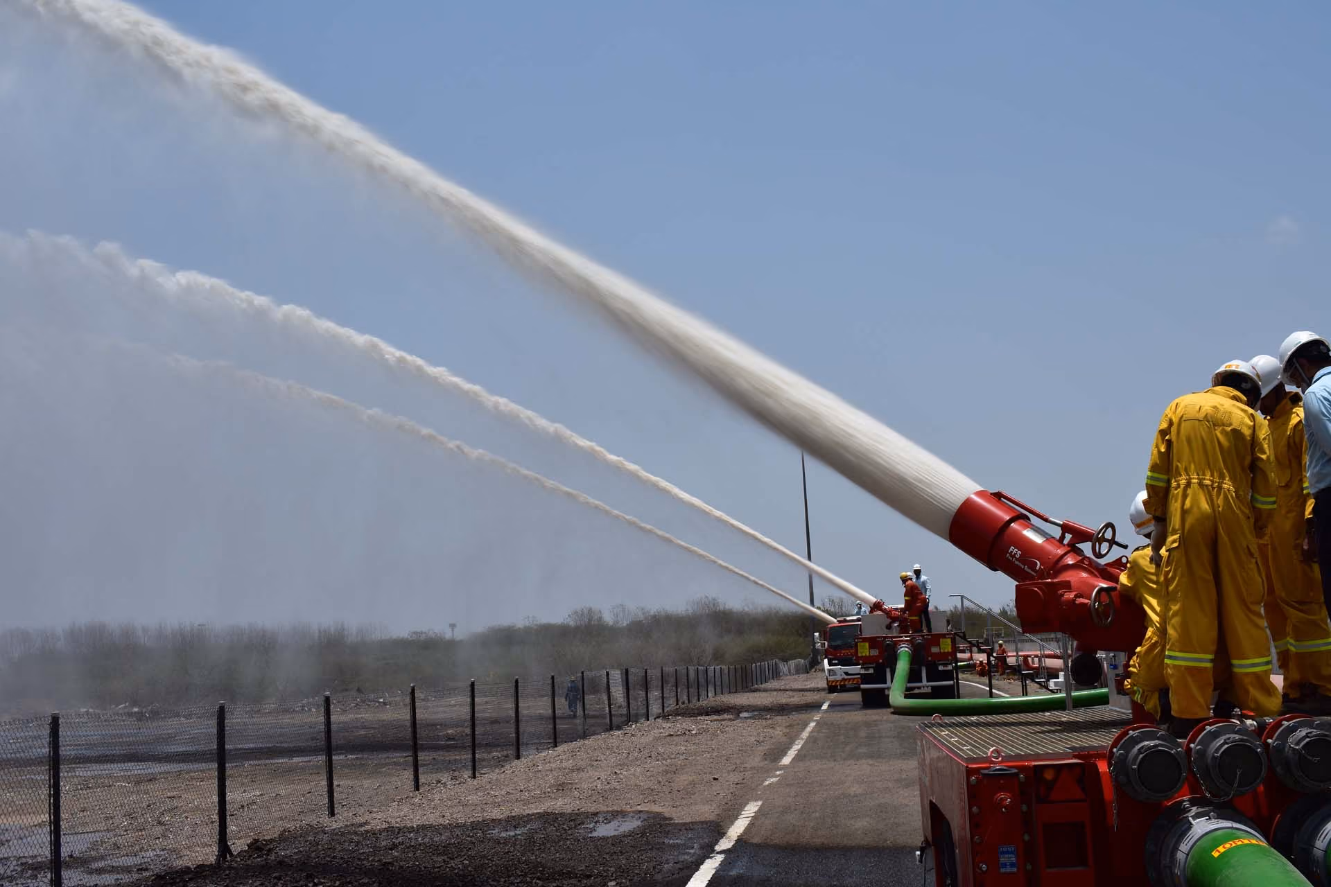 Firefighters in yellow suits operate large red water cannons spraying water over a fenced area.