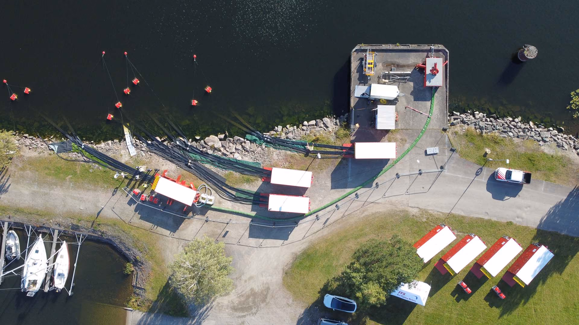 Aerial view of a water treatment or filtration site with tanks, pipes, boats, and vehicles near a body of water.
