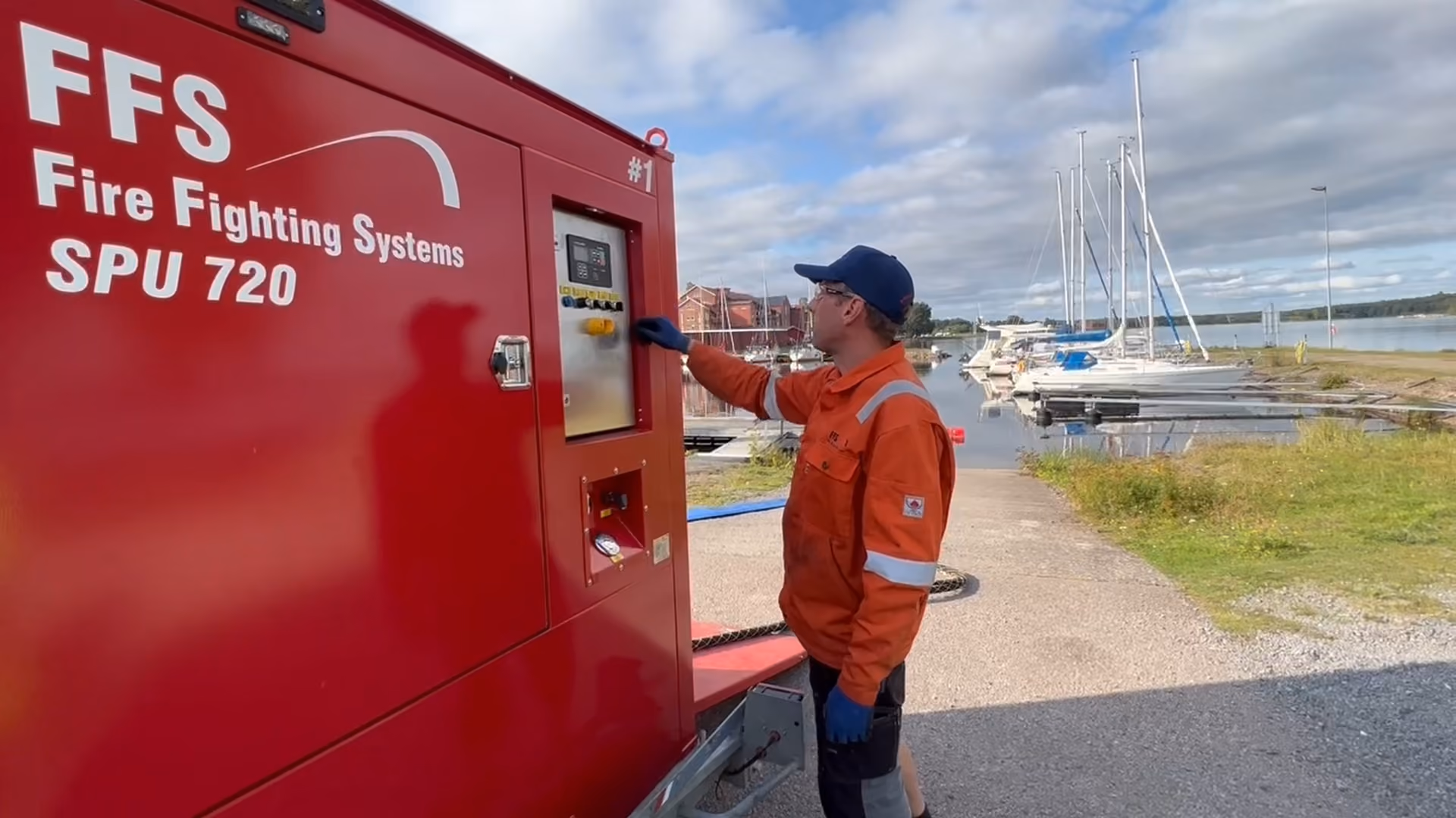 Worker in orange gear operates a red Fire Fighting Systems SPU 720 unit near a marina with sailboats.