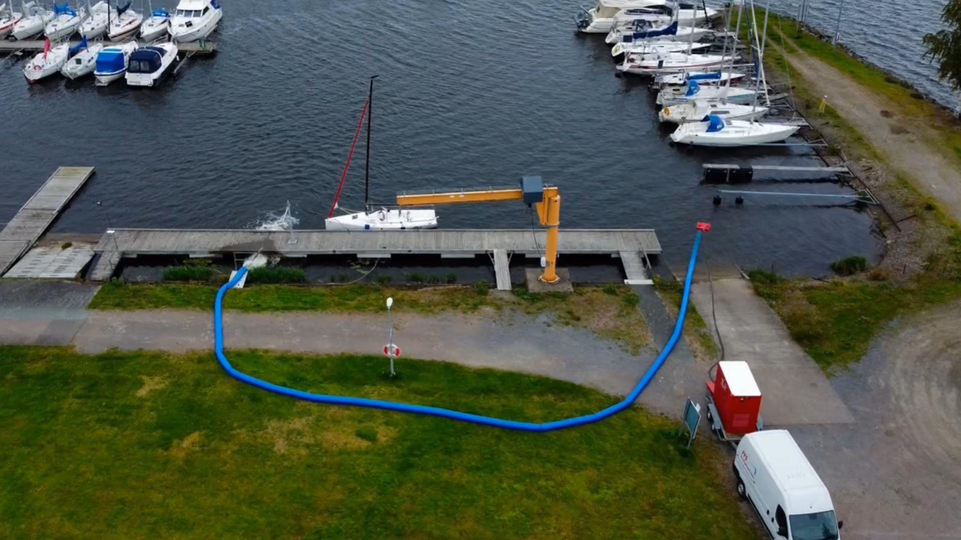 Small marina with docked sailboats and a white sailboat near a yellow crane on the dock.