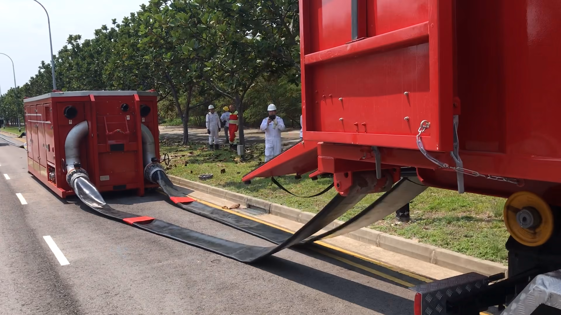 Large red industrial machine connected by wide black hoses on a road with workers in safety gear nearby.