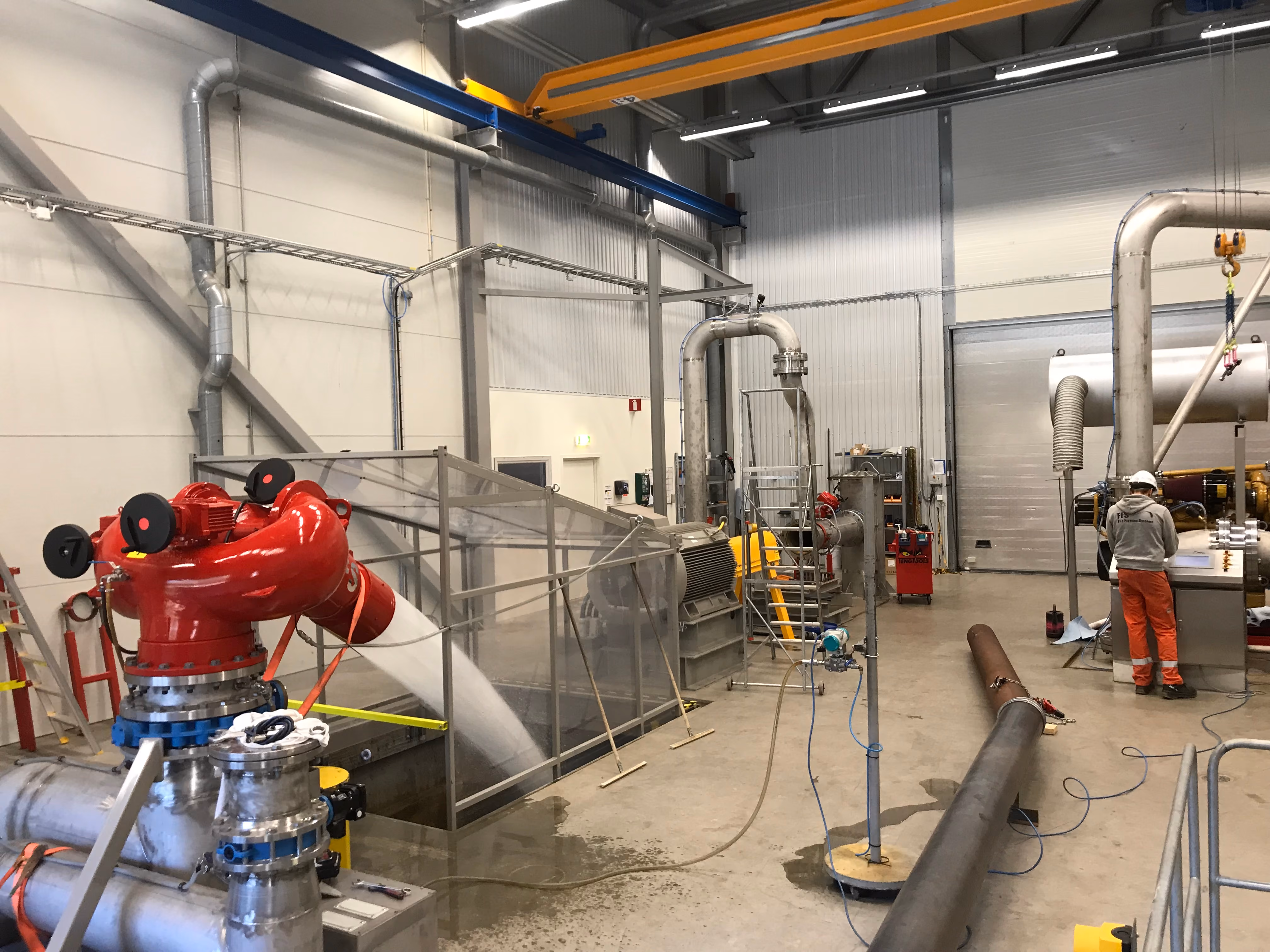 Industrial facility interior with pipes, a large red valve, flowing water, and a worker in safety gear.