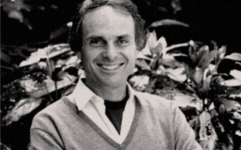 A black and white photograph of a smiling man with light skin and short dark hair. He stands in front of trees. 