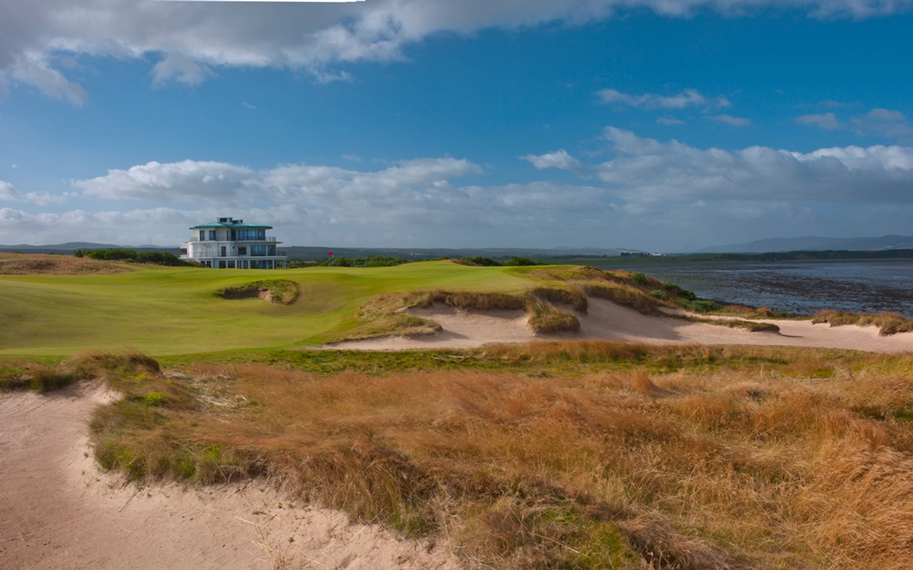 Golf course with sand bunkers, brown grass, and a clubhouse near a body of water under a partly cloudy blue sky.