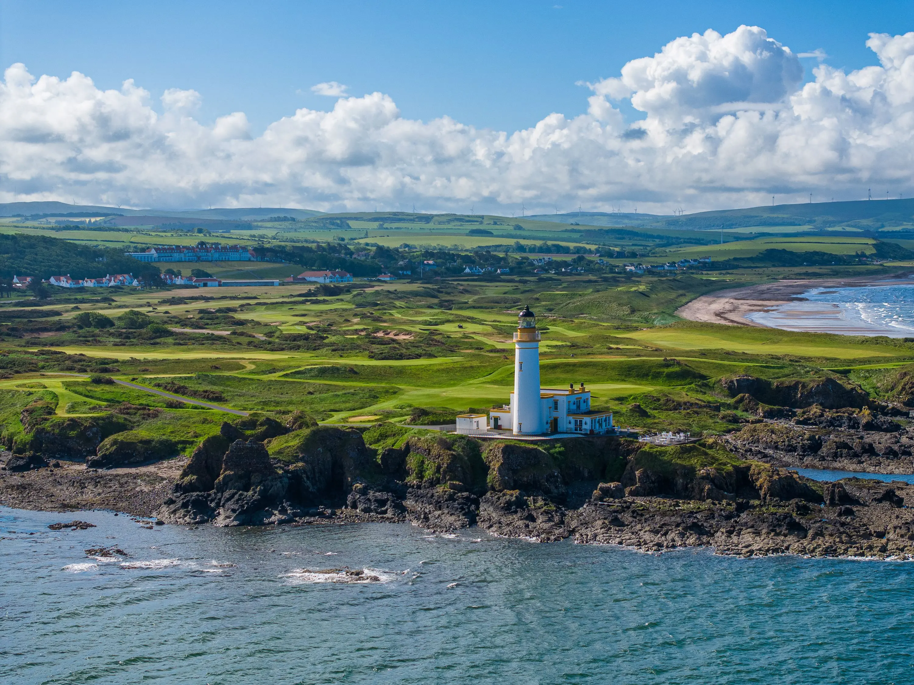 Lighthouse on rocky coast overlooking green fields and a distant seaside village under a partly cloudy blue sky.
