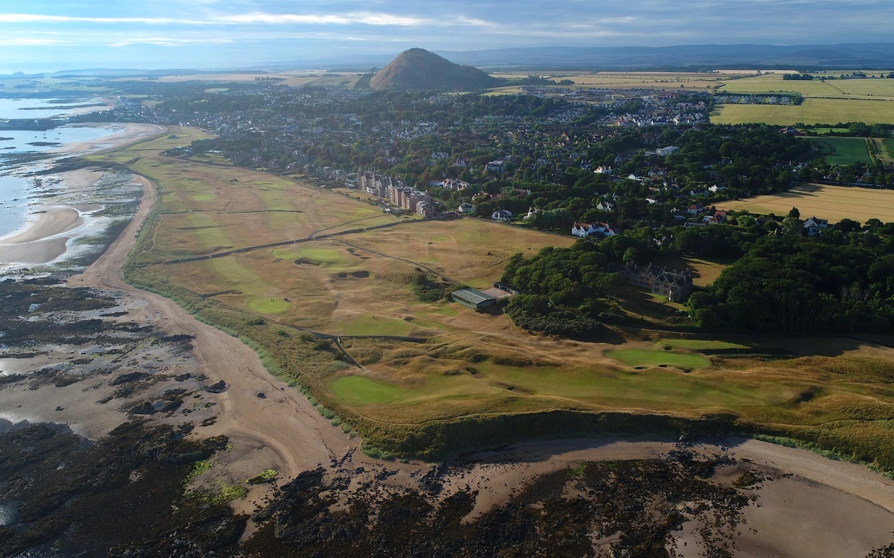 Aerial view of a coastal town with a sandy beach, golf course, and a large hill in the background under a cloudy sky.