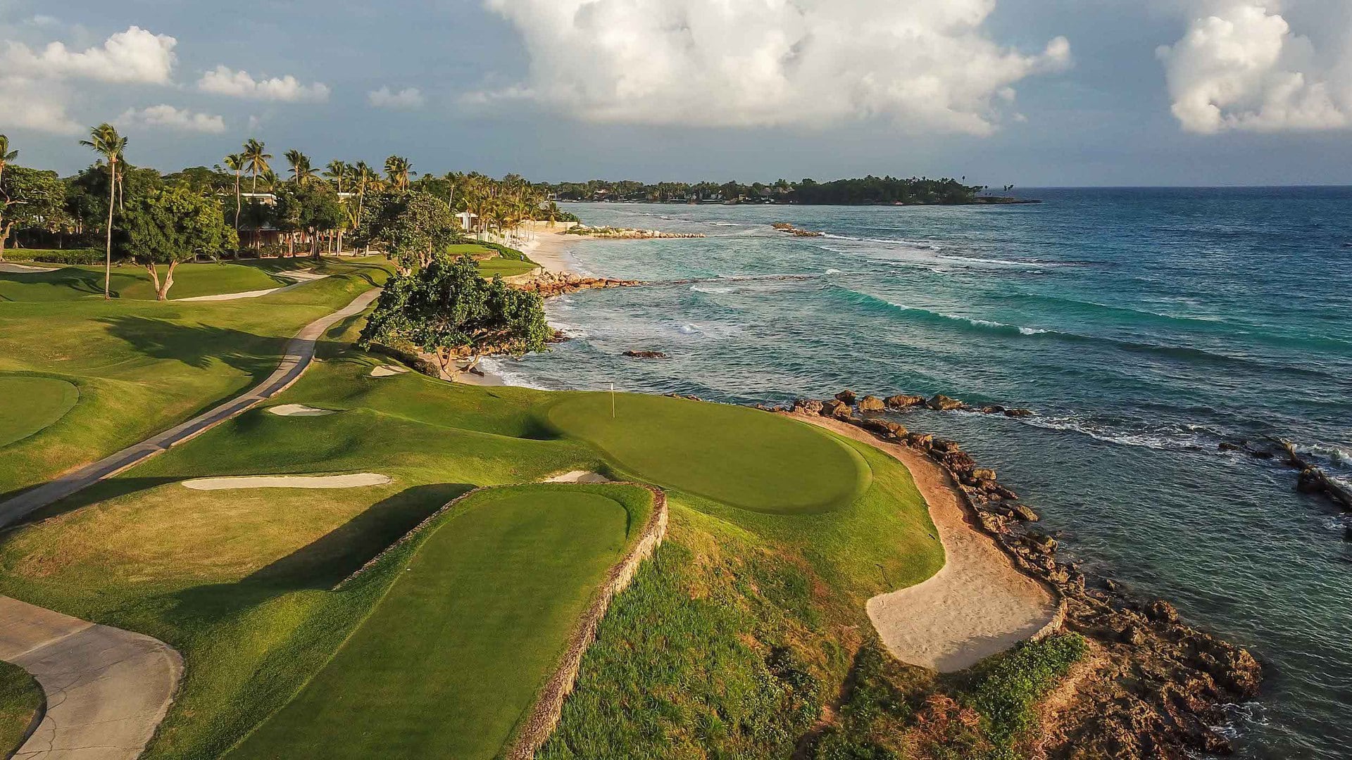 A coastal golf course with green fairways next to a sandy beach and turquoise ocean under a partly cloudy sky.
