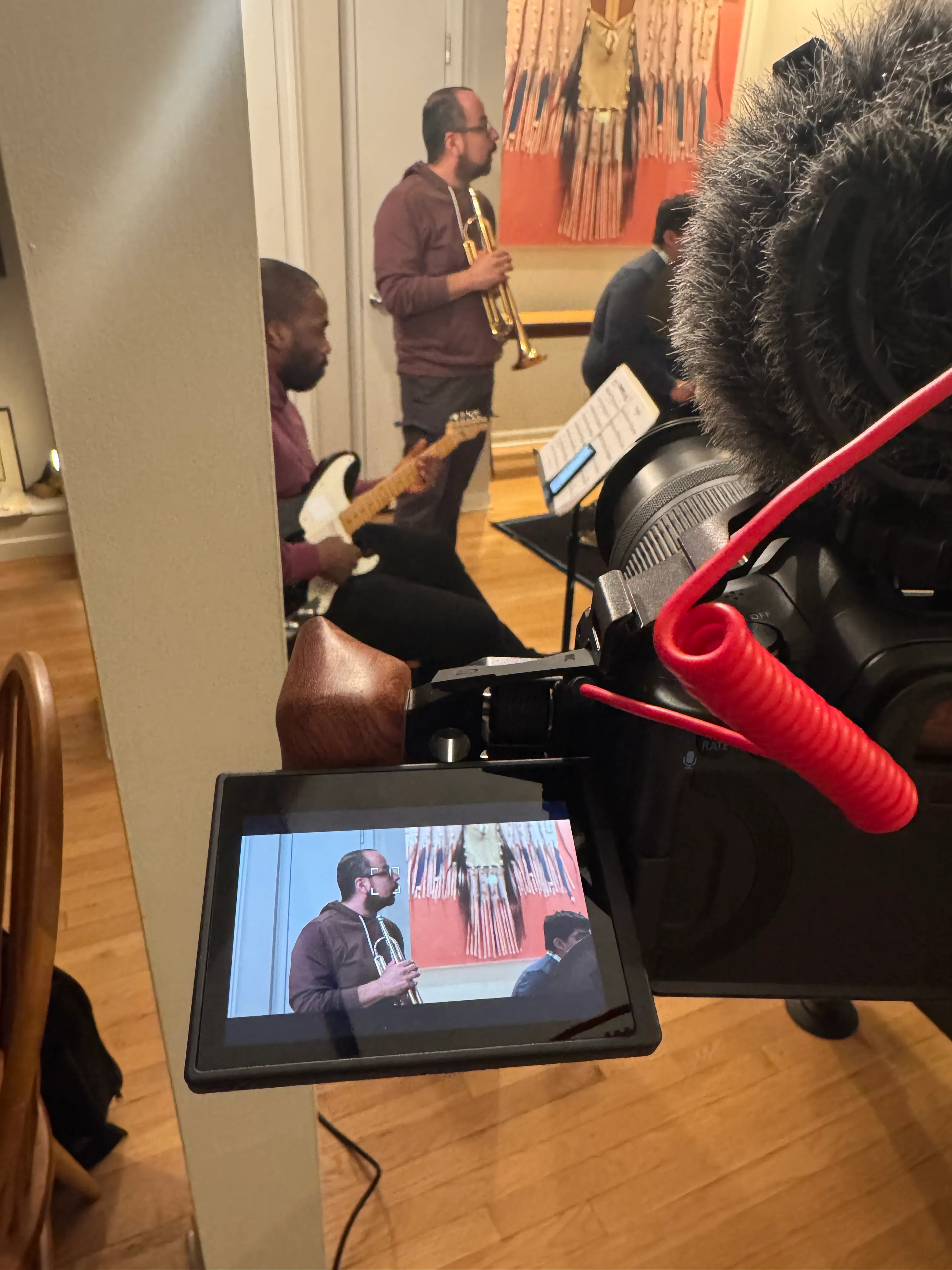 A camera monitor showing a music teacher being filmed in class, with a trumpet and guitar nearby