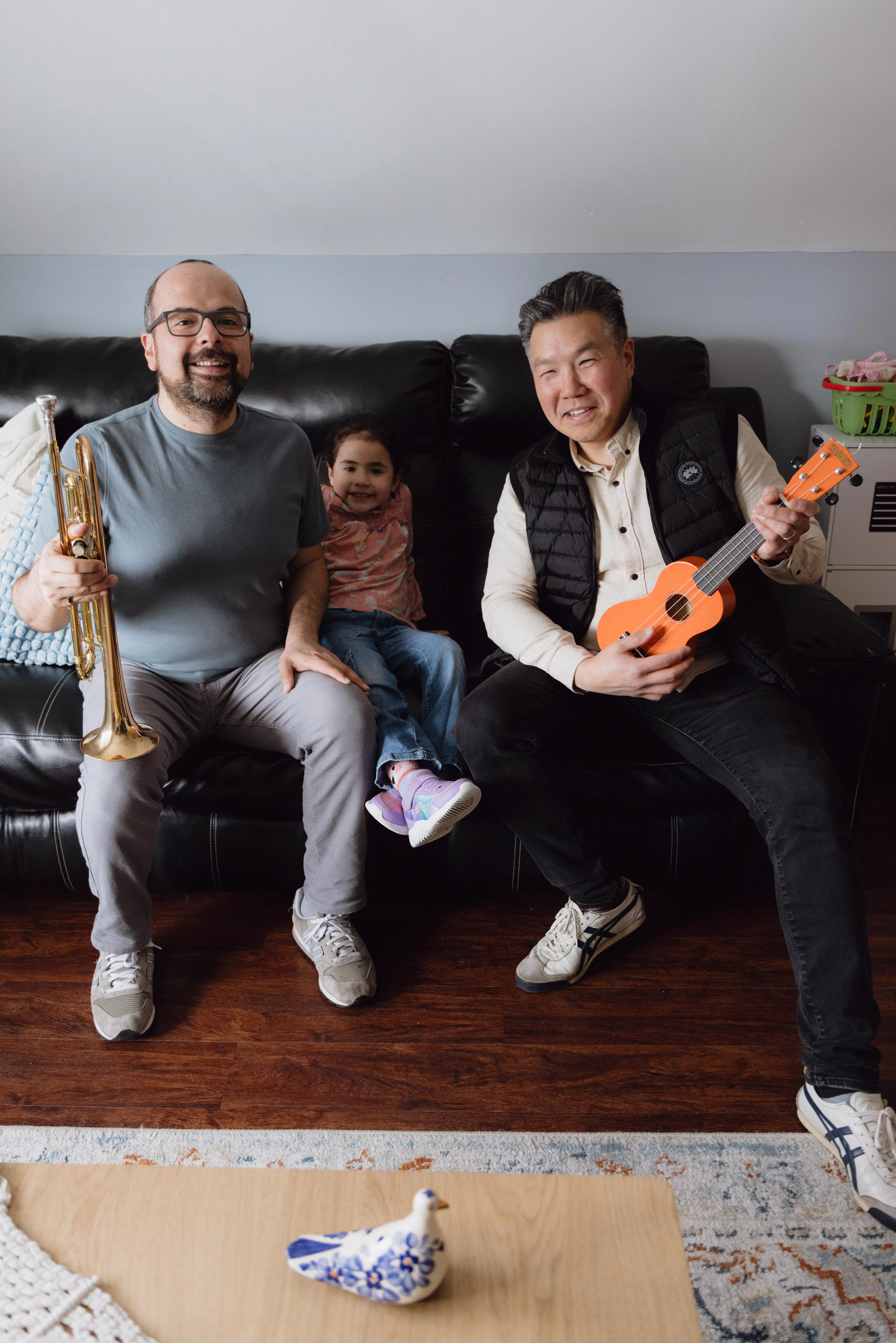 Two men sitting on a living room couch holding a trumpet and a ukulele, with a young child between them