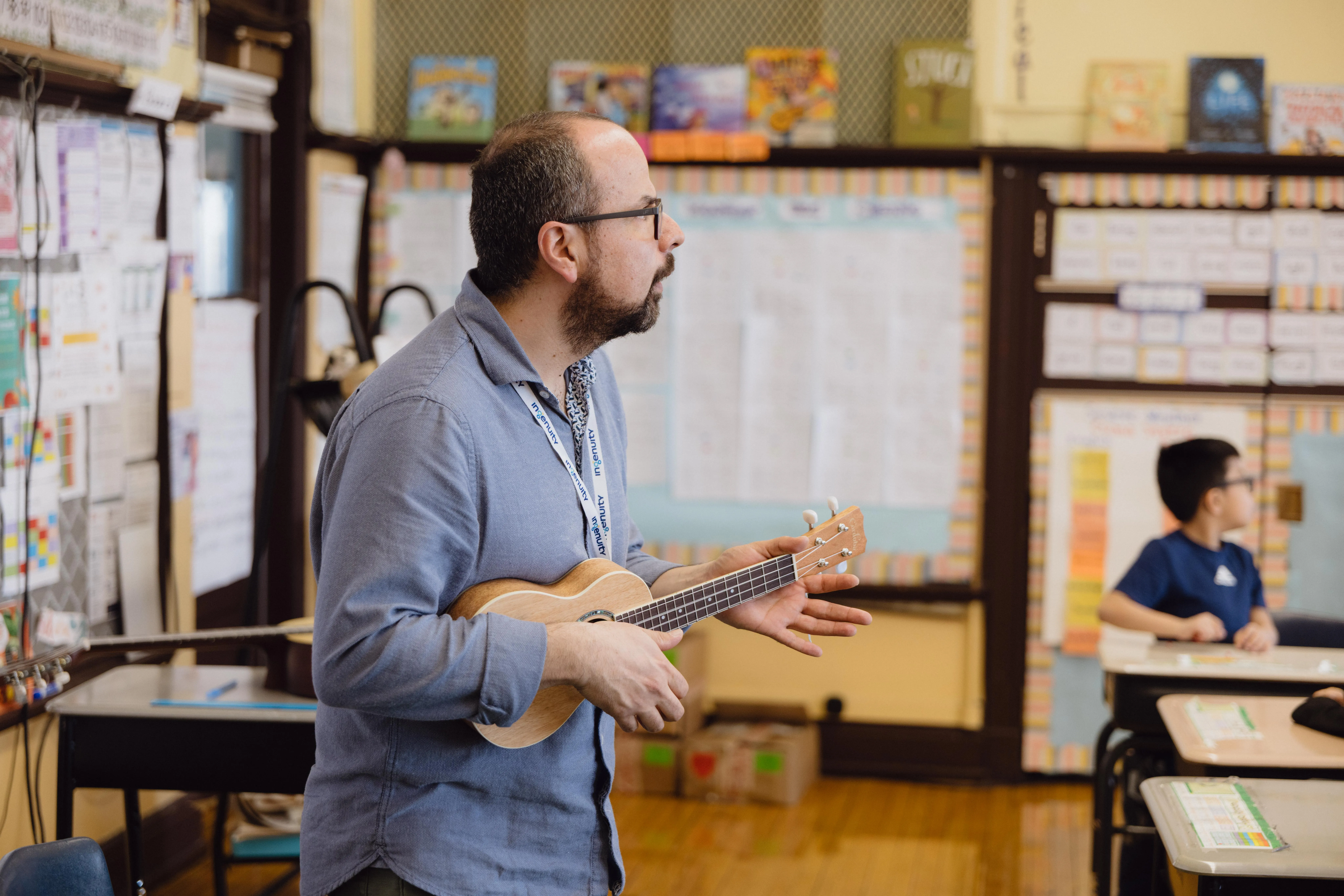 A teacher plays a ukulele at the front of his classroom while a student watches from a desk