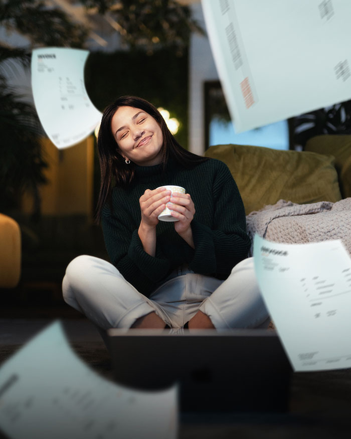 Tyytyväinen nuori nainen istuu tietokoneen edessä ja paperit lentävät hänen ympärillään