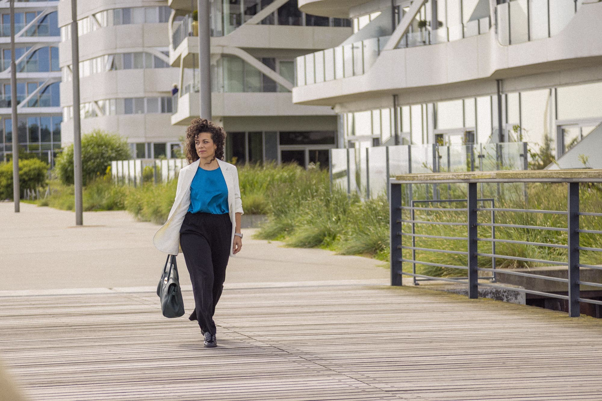 A woman walking along a modern boardwalk in an urban area, carrying a handbag, with contemporary white buildings and greenery in the background.