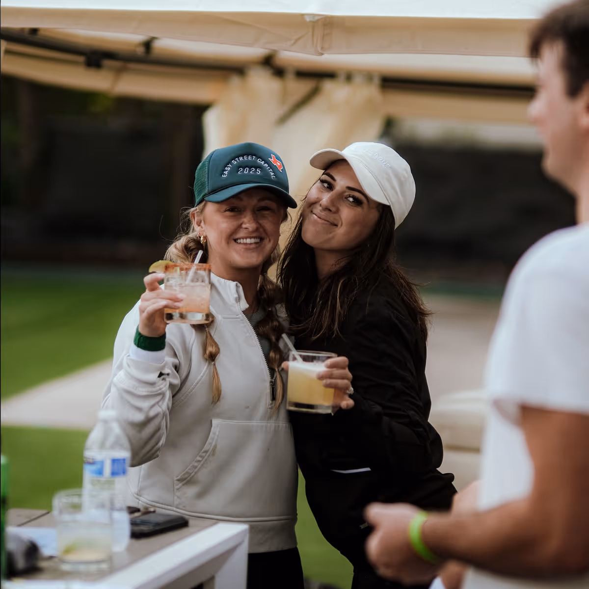 Two women smiling and holding drinks outdoors, one wearing a dark cap and the other a white cap.