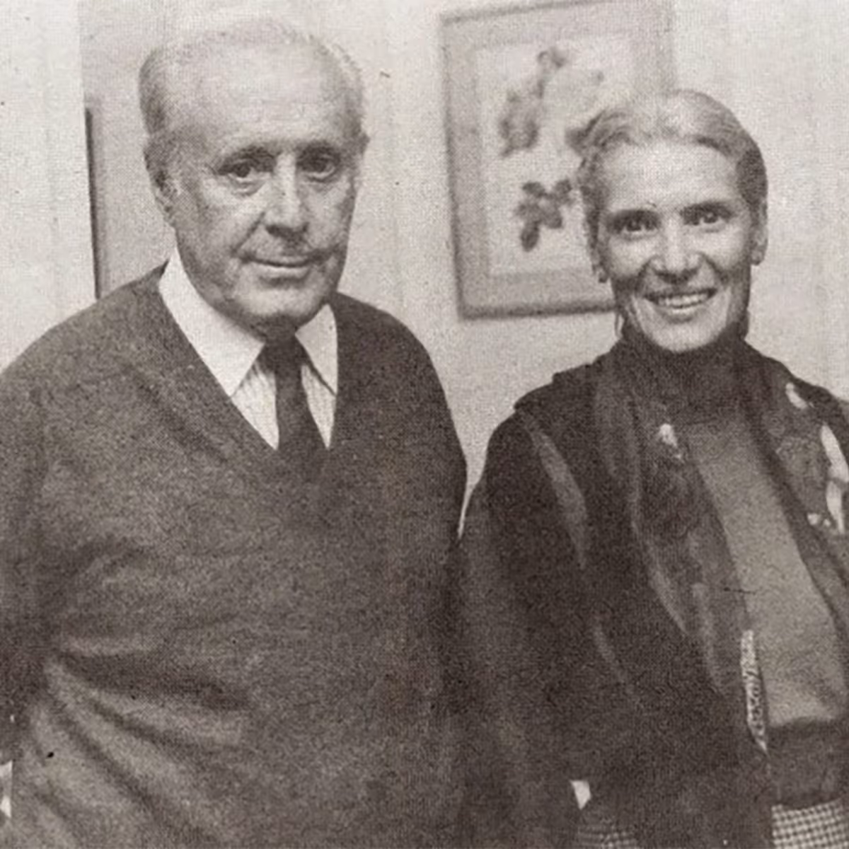 Black and white photo of an older man in a sweater and tie standing next to a smiling older woman with a scarf.