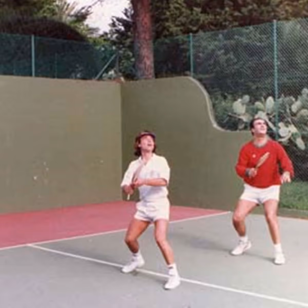 Two men playing padel on an outdoor court with green walls and chain-link fencing.