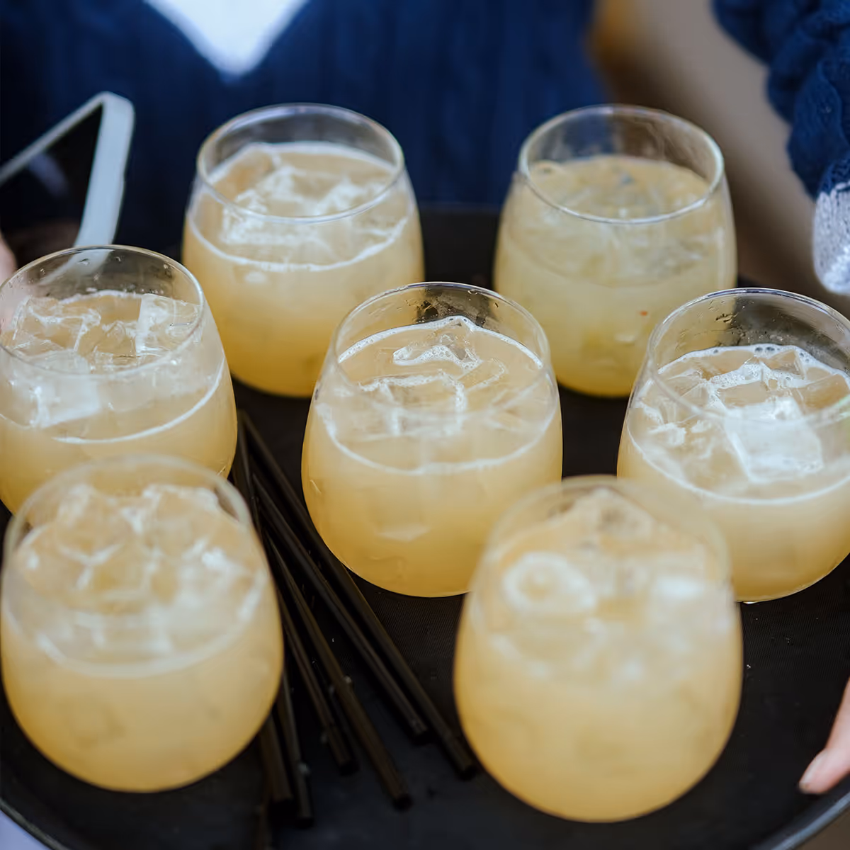 Tray of seven glasses filled with iced yellow-orange cocktails and black straws.
