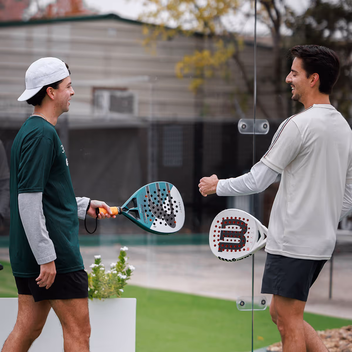 Two men smiling and holding padel rackets while standing on opposite sides of a glass wall at a padel court.