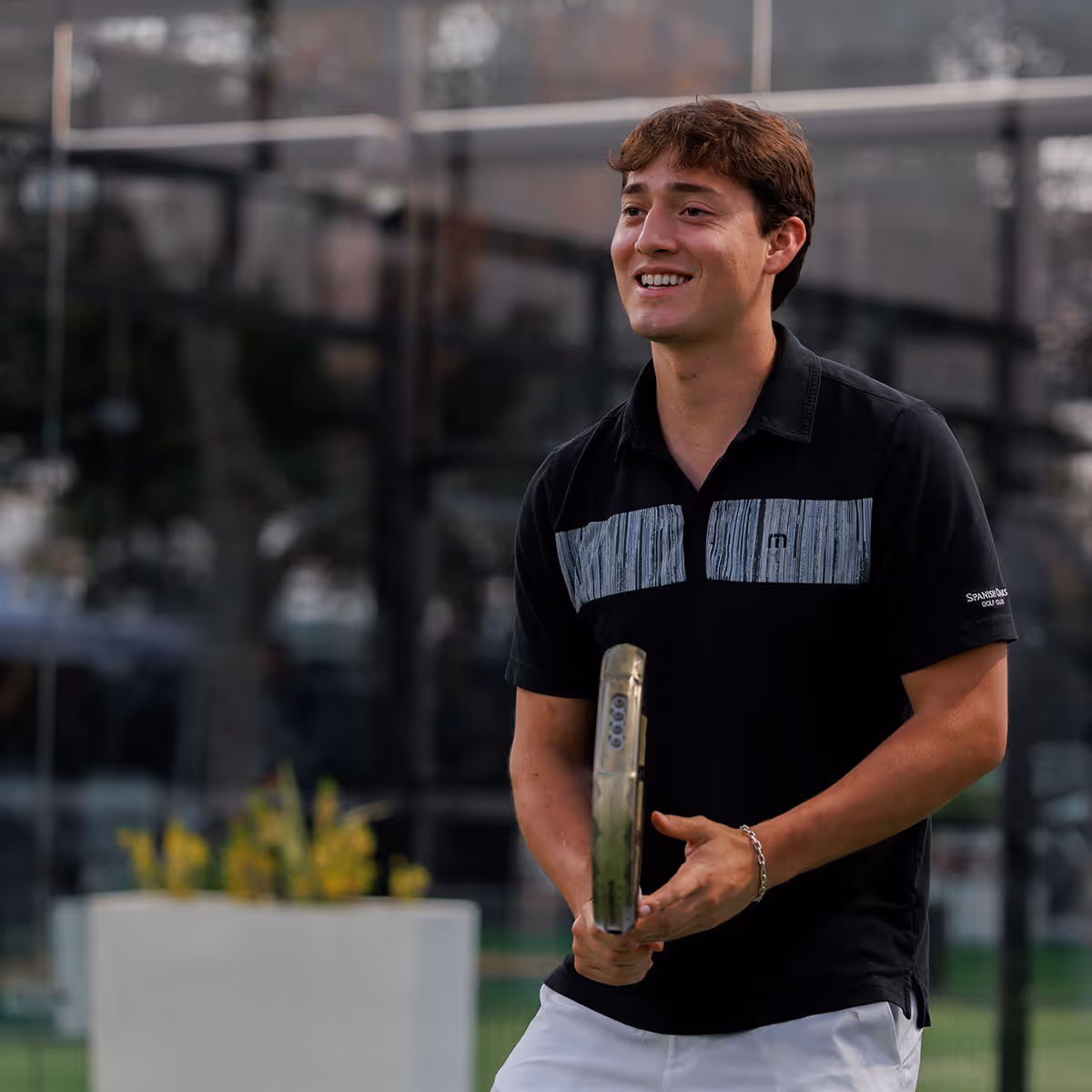Smiling young man holding a padel racket on an outdoor padel court.