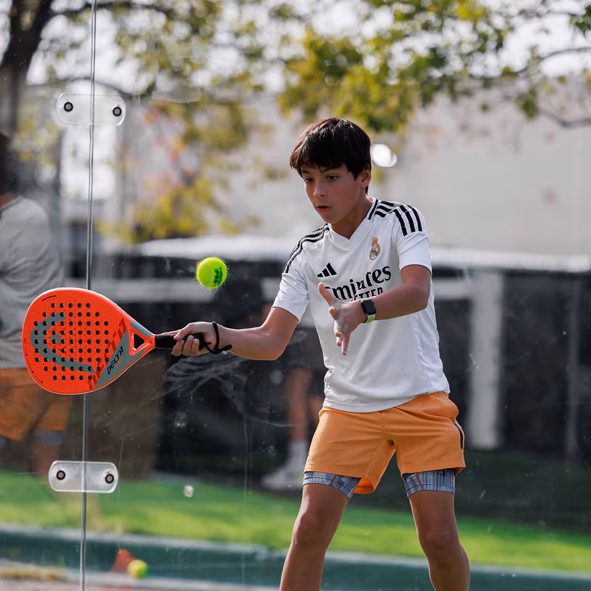Boy playing padel with orange racket hitting a yellow ball on a glass court.