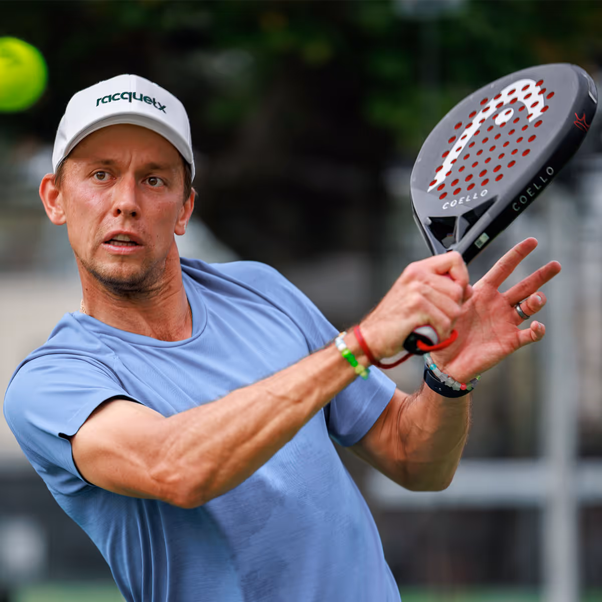 Man in a light blue shirt and white cap playing padel, swinging a black Coello racket.