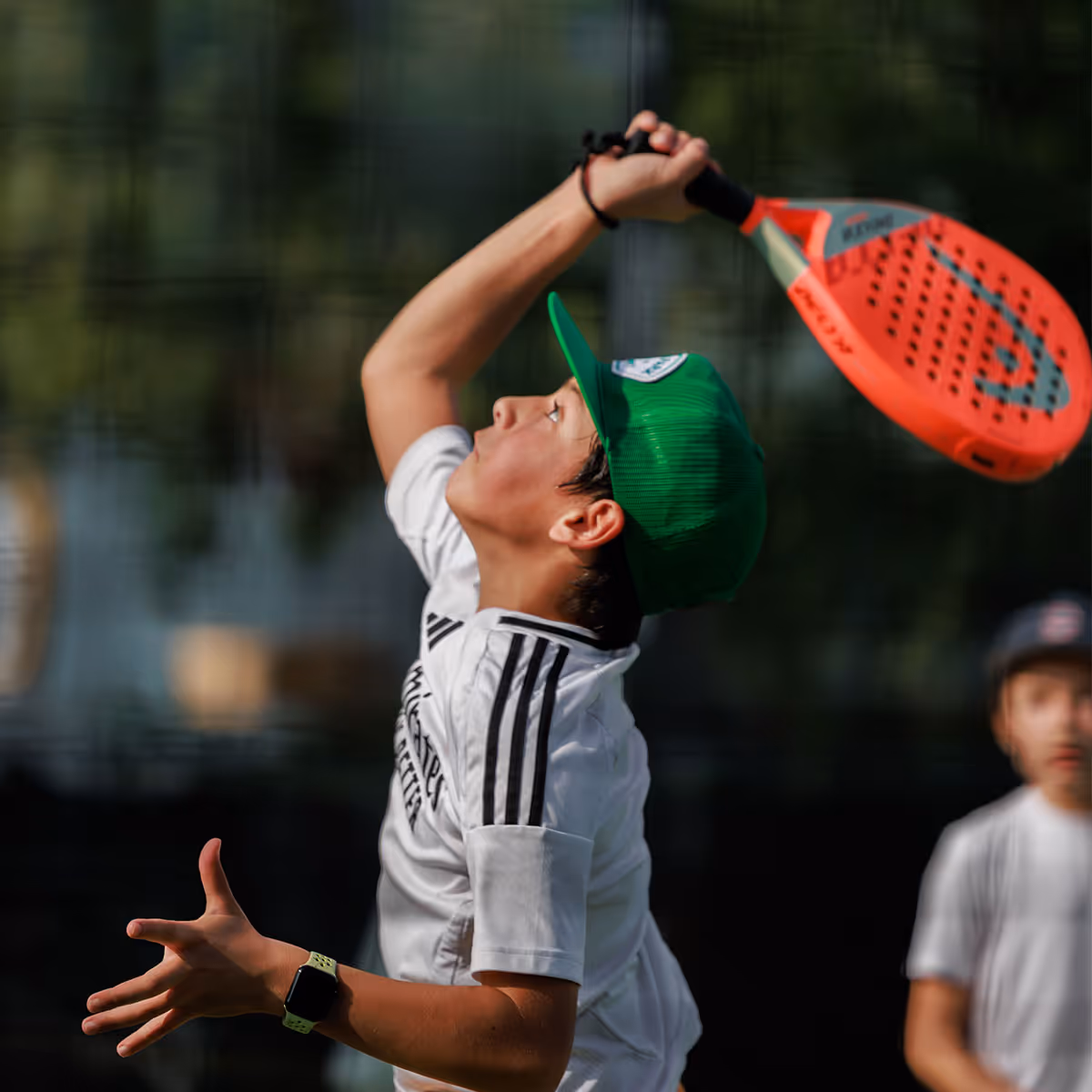 Young boy in a green cap and white sports shirt preparing to hit a ball with an orange padel racket during an outdoor game.