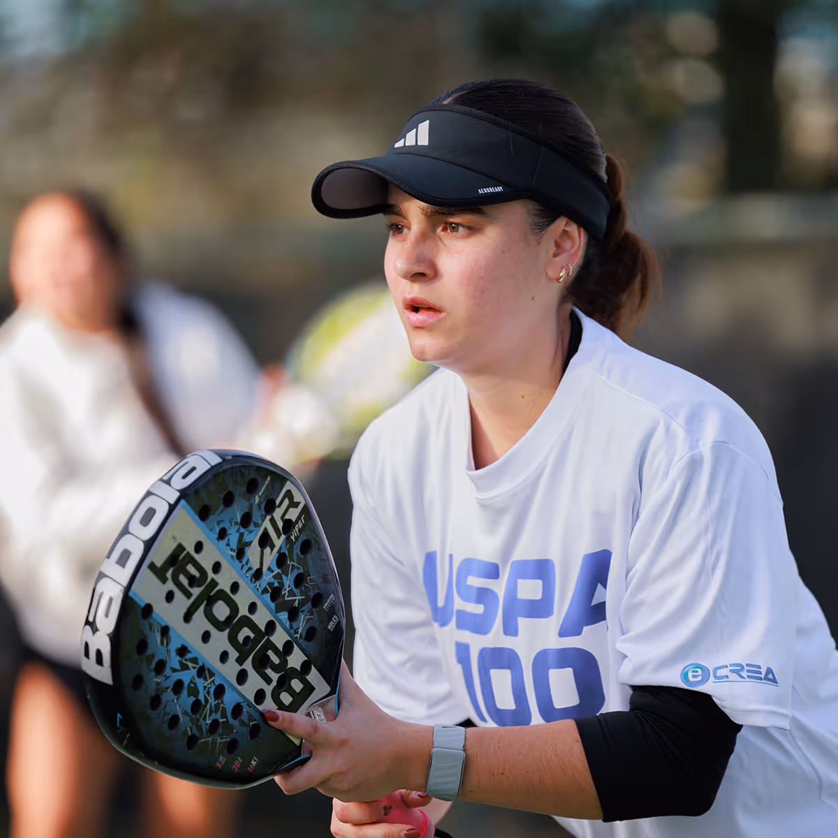 Female player wearing a black visor and white shirt holding a Babolat padel racket, focused during a game.