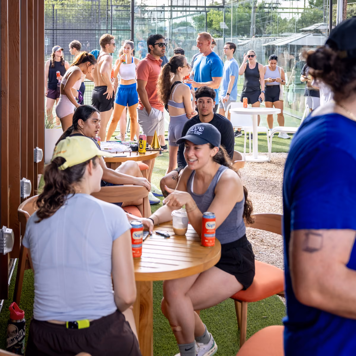 Two women smiling and holding drinks outdoors, one wearing a dark cap and the other a white cap.