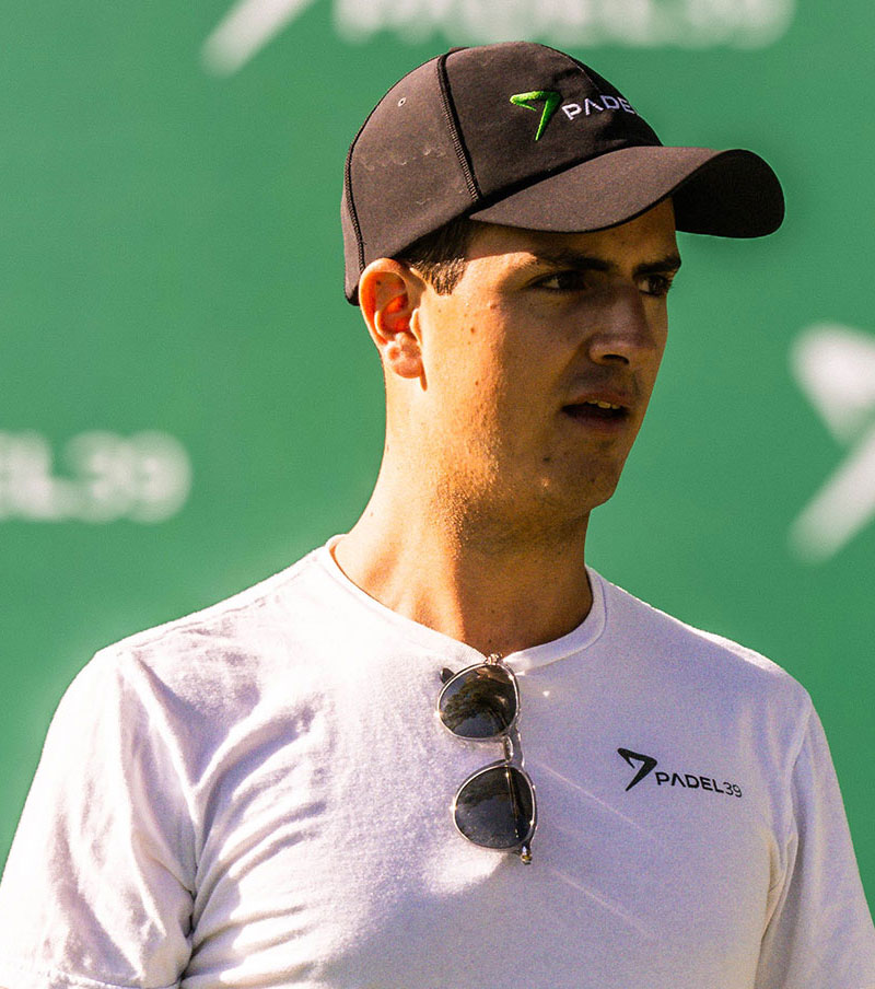 Man wearing white sportswear and a cap walking on a padel court with a racket bag over his shoulder.
