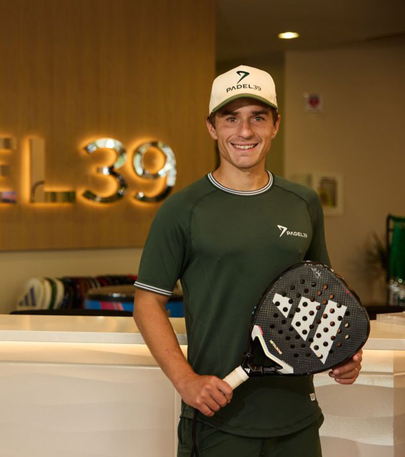 Man wearing white sportswear and a cap walking on a padel court with a racket bag over his shoulder.