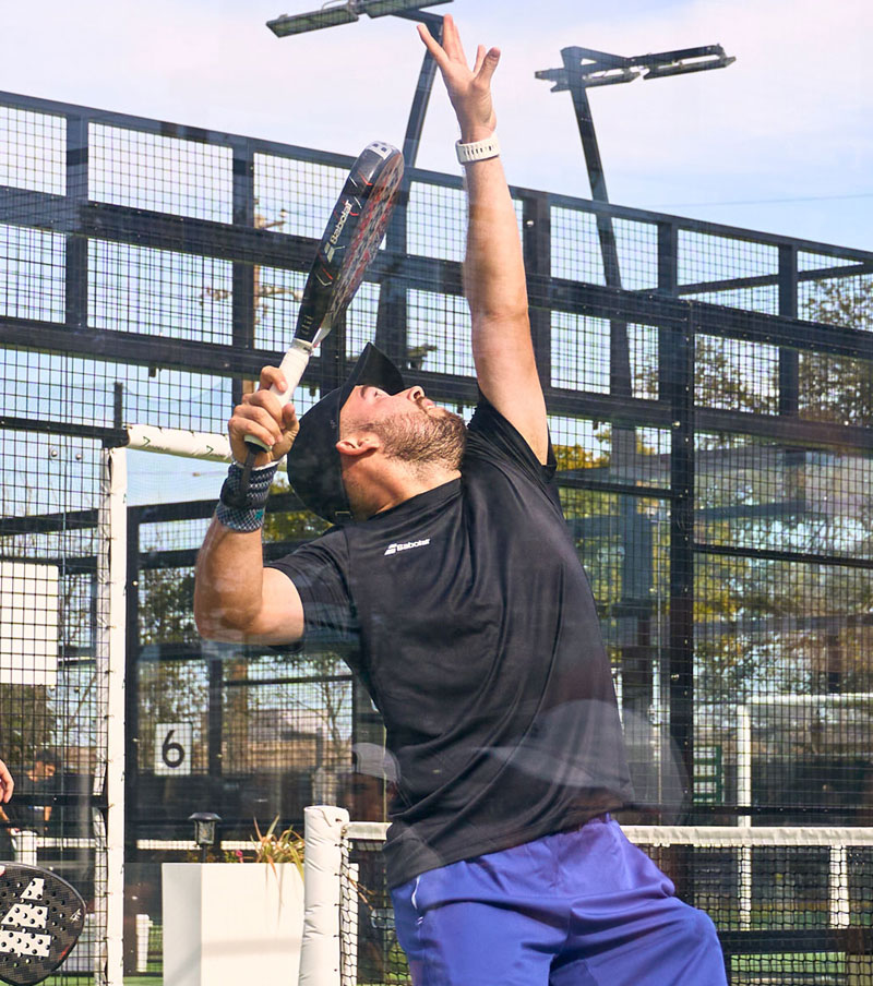 Man wearing white sportswear and a cap walking on a padel court with a racket bag over his shoulder.