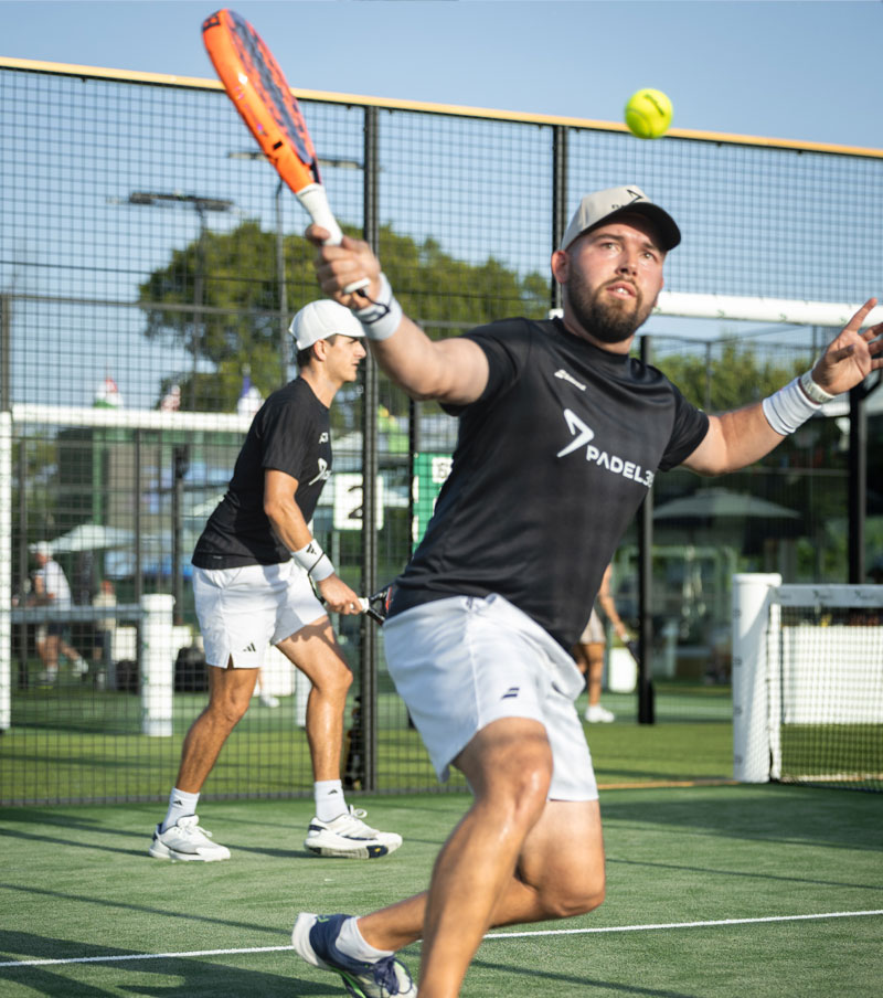 Man wearing white sportswear and a cap walking on a padel court with a racket bag over his shoulder.