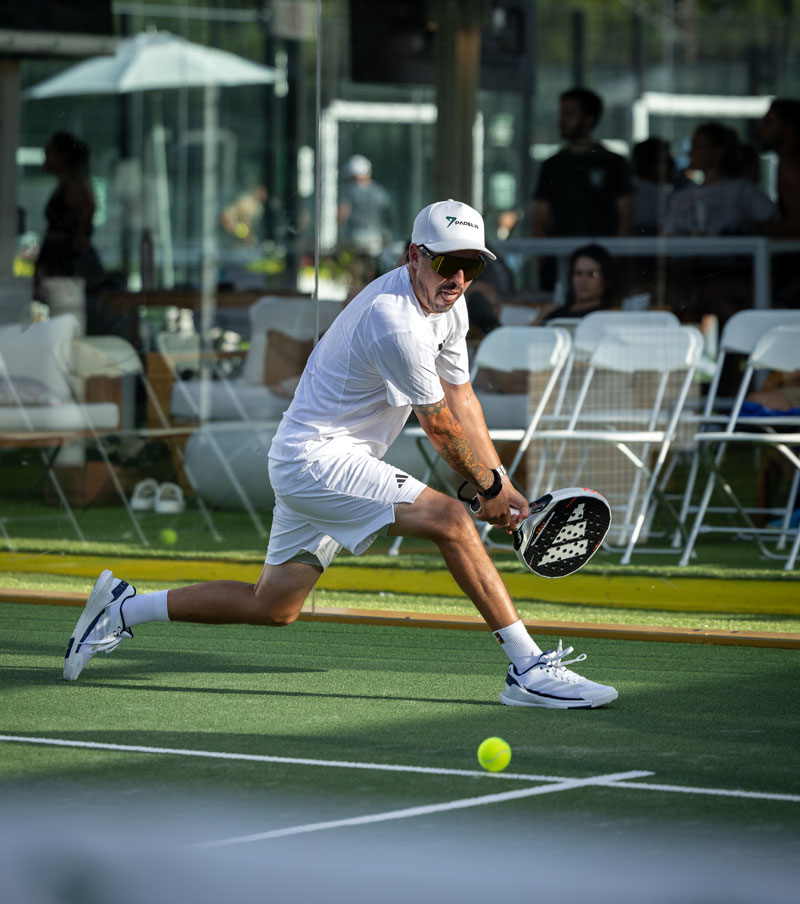 Man wearing white sportswear and a cap walking on a padel court with a racket bag over his shoulder.