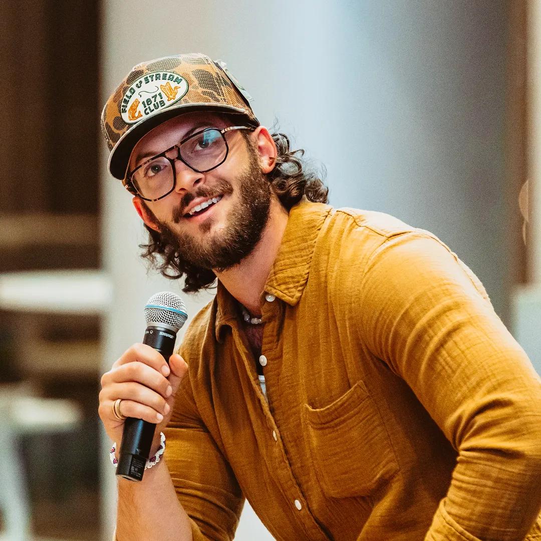 Smiling man with glasses, beard, and curly hair wearing a camo Field & Stream cap and mustard shirt speaking into a microphone.