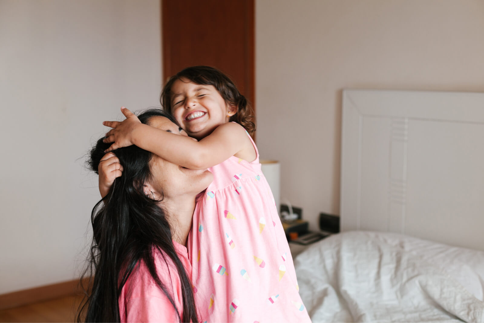 Positive mother and little daughter hugging on comfortable bed and smiling while resting in hotel room together