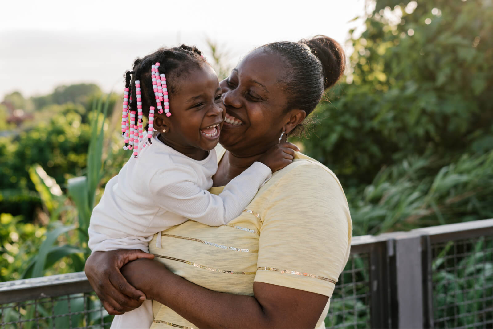 Mother smiling holding her daughter in her arms as she stands in a beautiful garden at sunset.