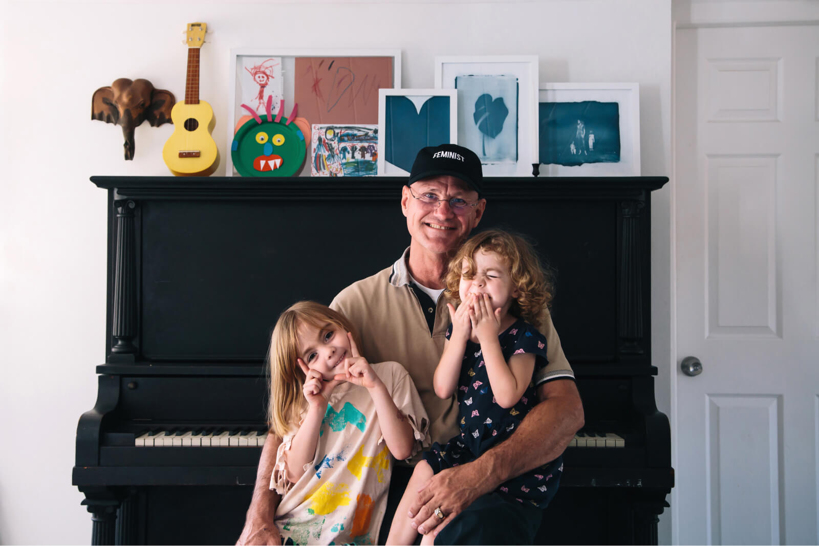 A grandpa wearing a hat, sits on a piano bench with his granddaughters