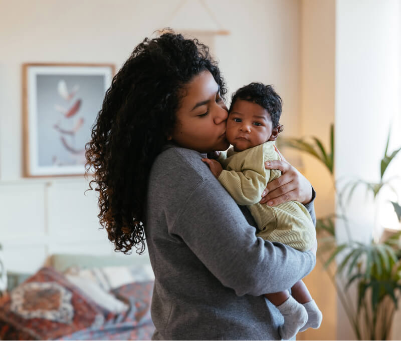 Side view of tender African American mother kissing sweet newborn on cheek while standing in cozy room at home