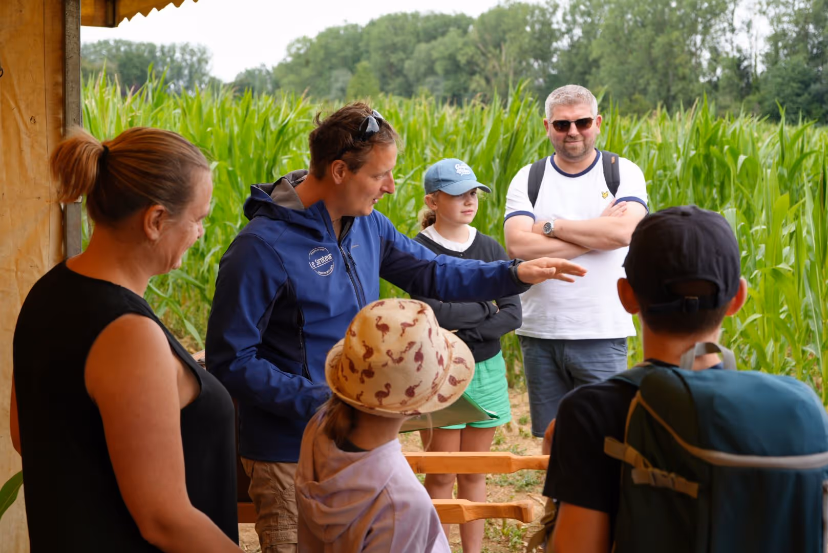 Un homme parle à un groupe de personnes dont des enfants devant un champ de maïs vert.