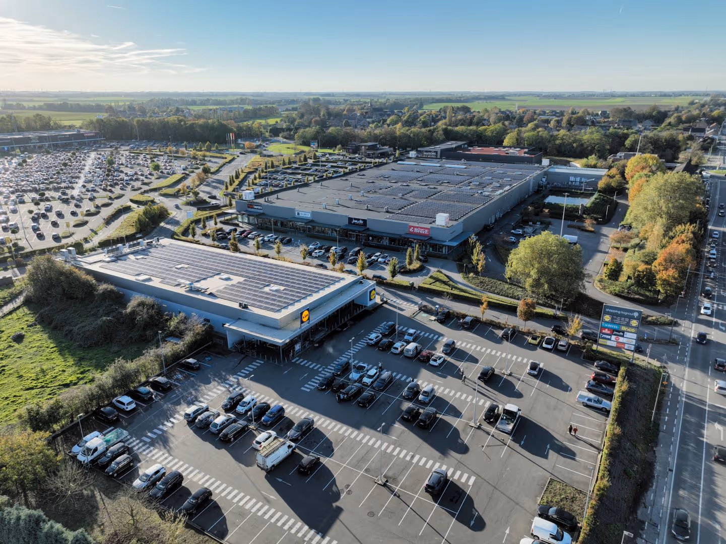 Vue aérienne d'un centre commercial avec parkings, bâtiments équipés de panneaux solaires et voitures garées.