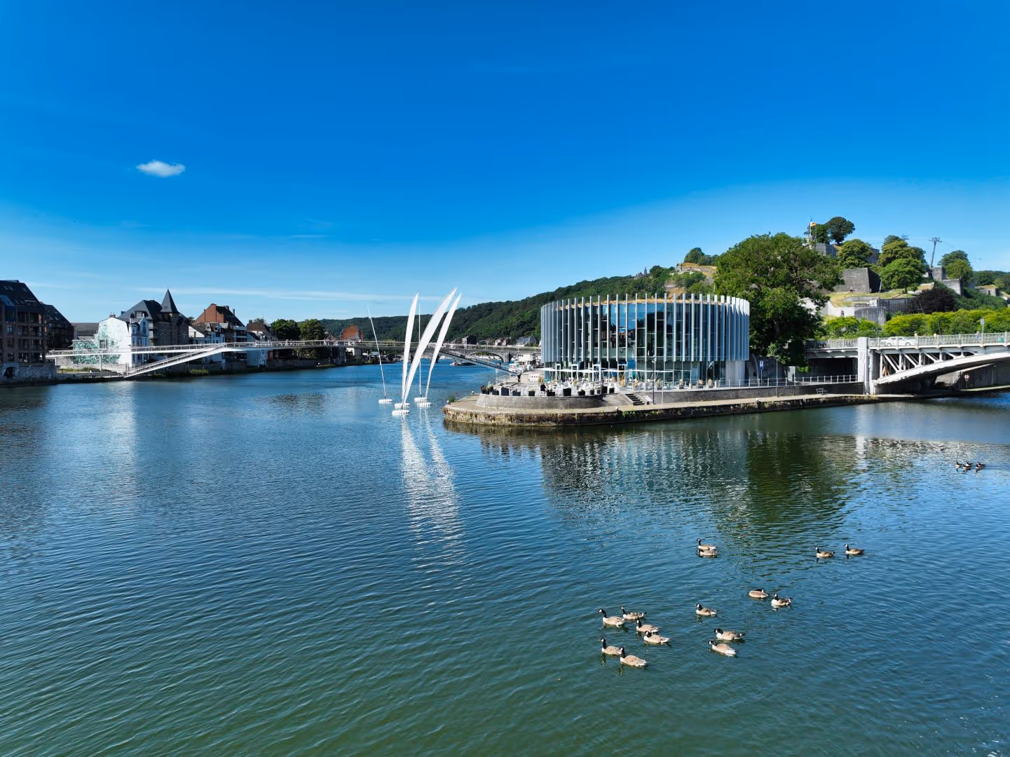 Bâtiment rond moderne au bord d'une rivière avec canards nageant sous un ciel bleu clair.