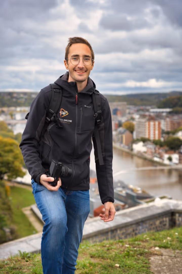 Un homme souriant tenant un appareil photo, avec un sac à dos, devant une rivière et une ville.
