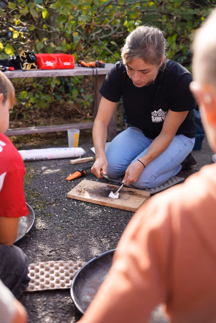 Une femme en pantalon bleu et t-shirt noir montre comment allumer un feu sur une planche en bois à l'extérieur.