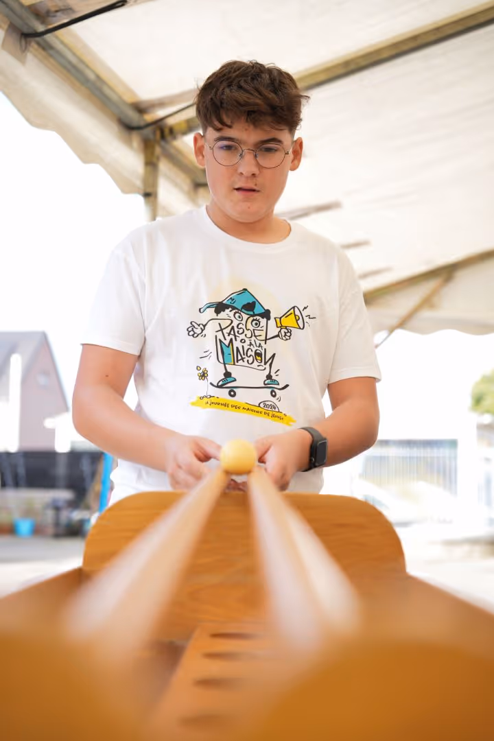 Un garçon portant des lunettes et un t-shirt blanc joue à un jeu de lancer de balle en bois sous un abri.