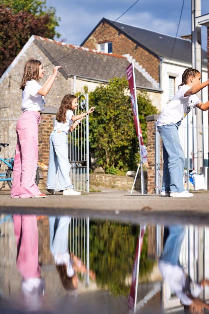 Trois enfants debout à l’extérieur, jouant et se reflétant dans une flaque d’eau sur le sol.